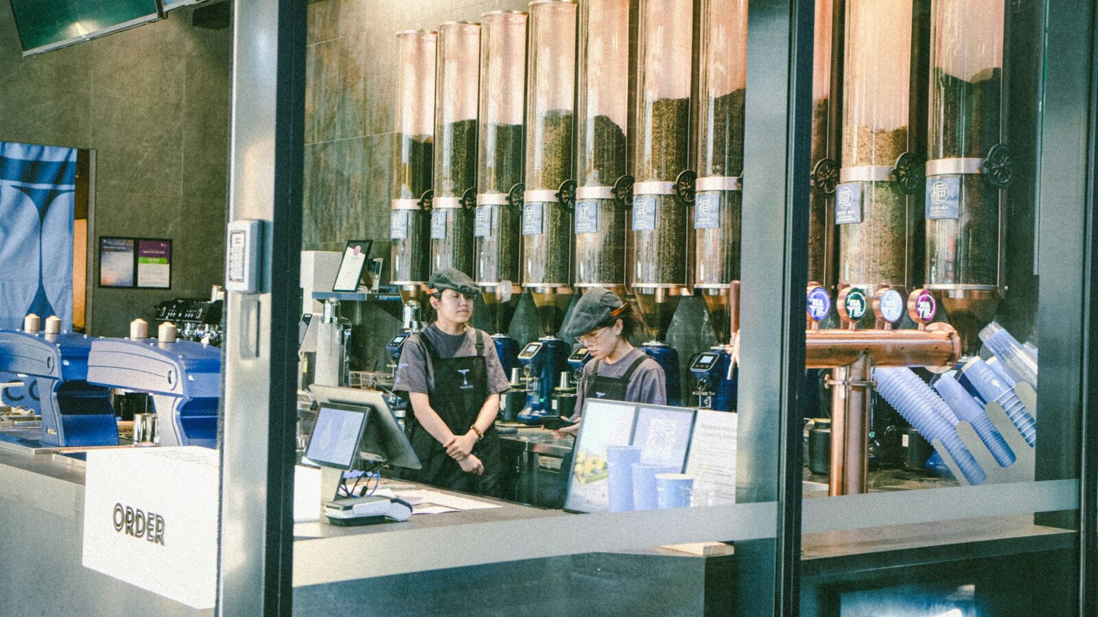Dois baristas atrás do balcão de uma cafeteria, vistos de quem está na fila.