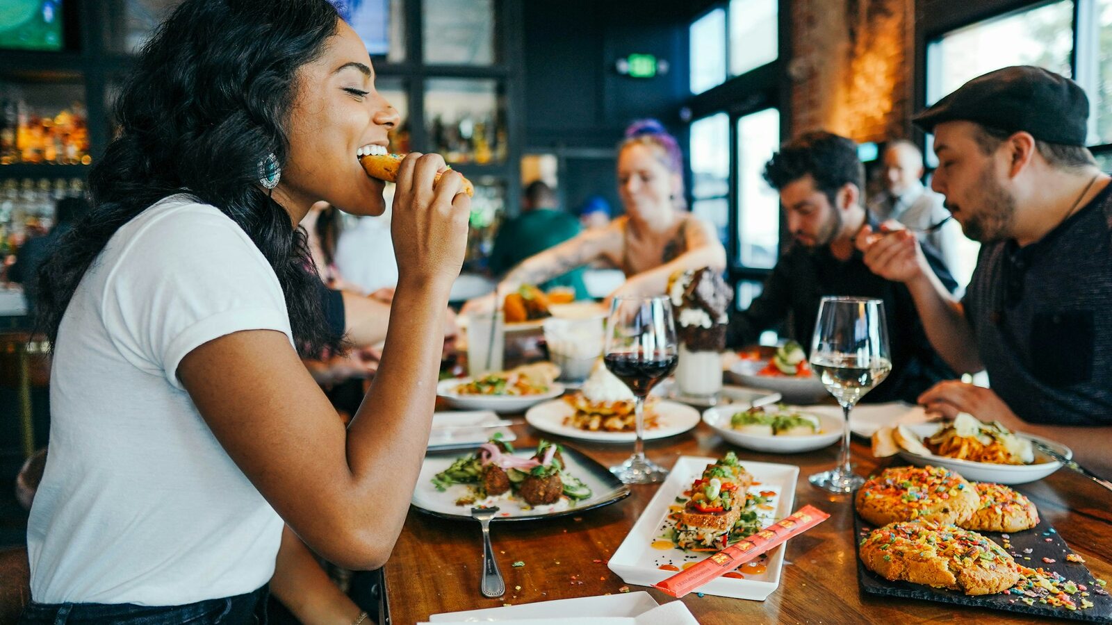 Mesa comprida de restaurante cheia no horário do almoço, com pratos servidos e clientes comendo.