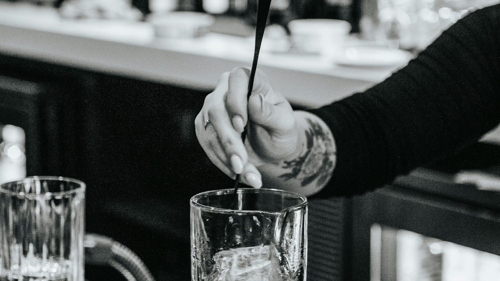 Bartender mexendo um drinque com gelo num copo, atrás do balcão.