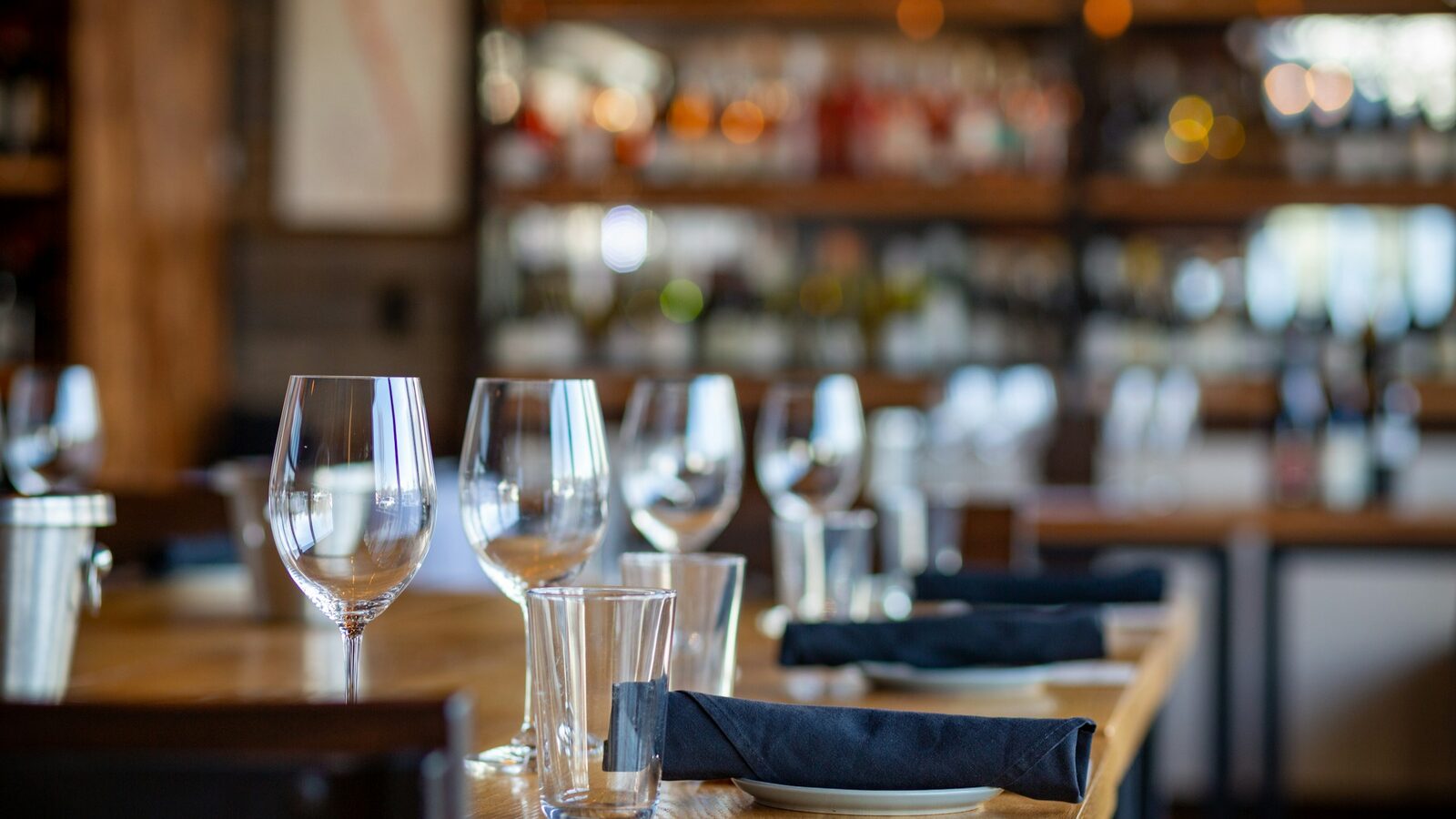 Mesa de restaurante posta com taças e copos altos, com o salão ao fundo.