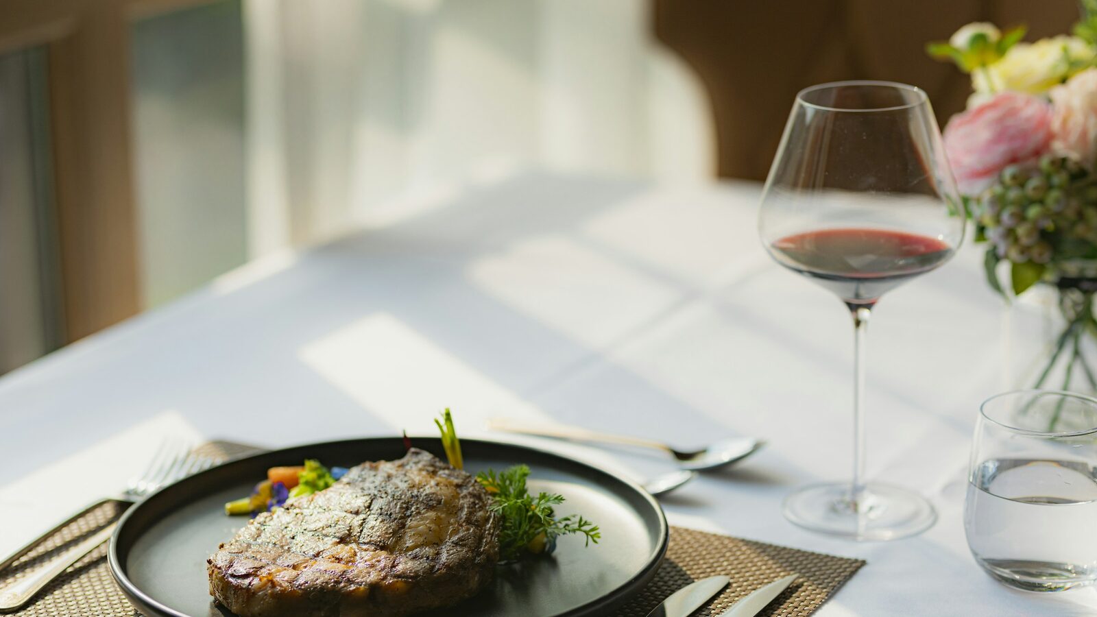 Bife servido num prato escuro, ao lado de uma taça de vinho tinto e um copo de água.